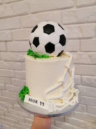 Tort z dekoracją piłkarską, zwieńczony piłką nożną, z napisem 'Igor 11' na podstawie, na tle białej ceglanej ściany.