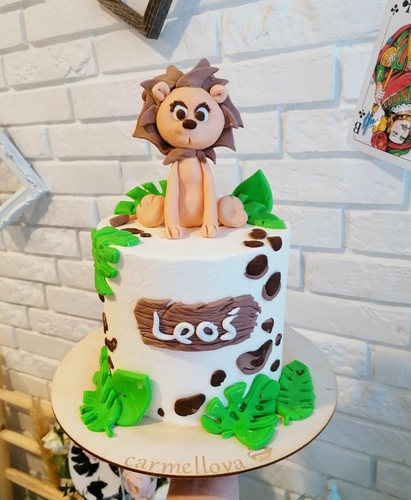 Tort urodzinowy z białym kremem, dekorowany brązowymi plamkami, zielonymi liśćmi i figurką lwa z modeliny na górze, z napisem 'Leoś' na brązowej tabliczce.