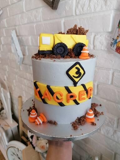 Tort urodzinowy w stylu budowlanym z żółtą ciężarówką na górze, dekorowany imieniem 'Kacper', cyfrą '3' i stożkami drogowymi na tle ceglanej ściany.
