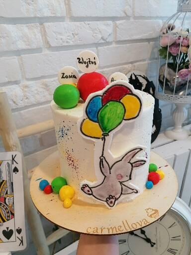 Tort urodzinowy z białym kremem, dekorowany rysunkiem królika trzymającego balony, z imionami Zosia i Wojtuś na małych tabliczkach, na drewnianej podstawie z logo 'carmellova'.