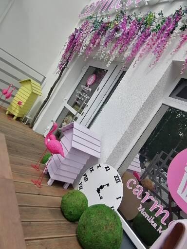 Wejście do lokalu z dekoracjami: różowe flamingi, budka dla ptaków, zegar, zielone kule i kwiaty nad drzwiami.