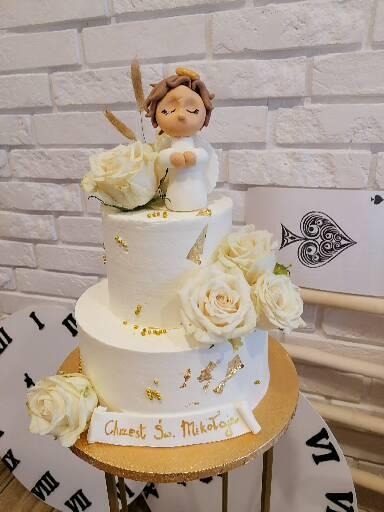 Dwupiętrowy tort z białego kremu, udekorowany białymi różami, złotymi płatkami i figurką anioła na szczycie, z napisem 'Chrzest Św. Mikołaja' na dolnym piętrze, umieszczony na złotym stojaku na tle...