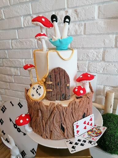 Fantazyjny tort inspirowany Alicją w Krainie Czarów: grzybki muchomory, nogi wystające z niebieskiej filiżanki, zegar z rzymskimi cyframi, drzwi do nory królika i karty do gry.