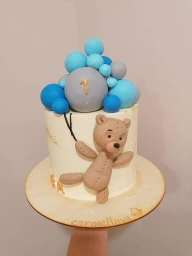 Tort urodzinowy dla roczniaka z figurką misia i niebieskimi balonami na białym tle.