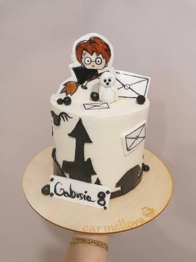 Tort urodzinowy z motywem Harry'ego Pottera, biały krem, czarne dekoracje, personalizowany napis Gabrysia 8 na drewnianej podstawie.
