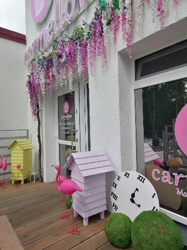 Fasada budynku z dekoracjami kwiatowymi, pastelowa budka, zegar i różowe flamingi na drewnianym podeście przed wejściem.