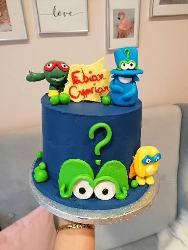 Niebieski tort urodzinowy z postaciami z bajek wykonanymi z lukru, z napisem 'Fabian Cyprian' na żółtym tle, ustawiony na srebrnej paterze.