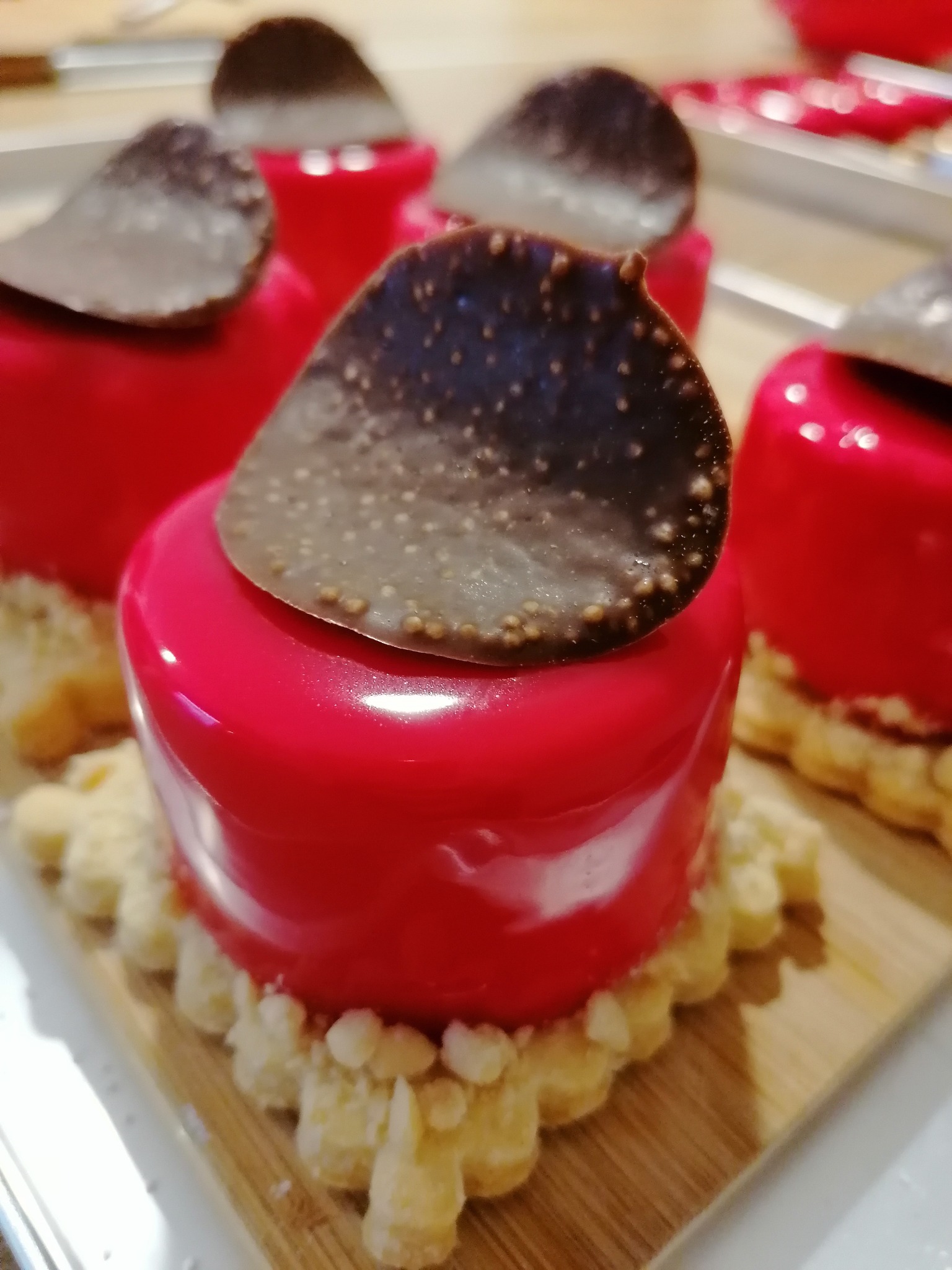 Mini deserki z błyszczącą, malinową glazurą i czekoladową dekoracją na kruchym spodzie, ułożone na drewnianej desce.