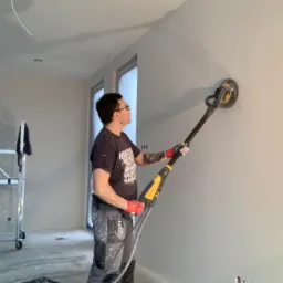 Pracownik w czerwonych rękawicach szlifuje ścianę szlifierką z długim uchwytem, widoczne ślady po obróbce, w tle rusztowanie, puszki z farbą Flűgger i narzędzia.