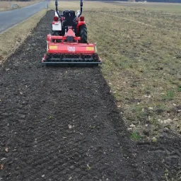 Czerwony traktor z glebogryzarką przygotowuje ziemię pod uprawę na polu przylegającym do drogi asfaltowej, w tle widoczny las.