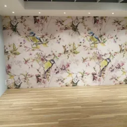 Wnętrze pokoju z drewnopodobną podłogą ułożoną z paneli i ścianą pokrytą tapetą z motywem ptaków i kwiatów, oświetlone punktowymi lampami sufitowymi.