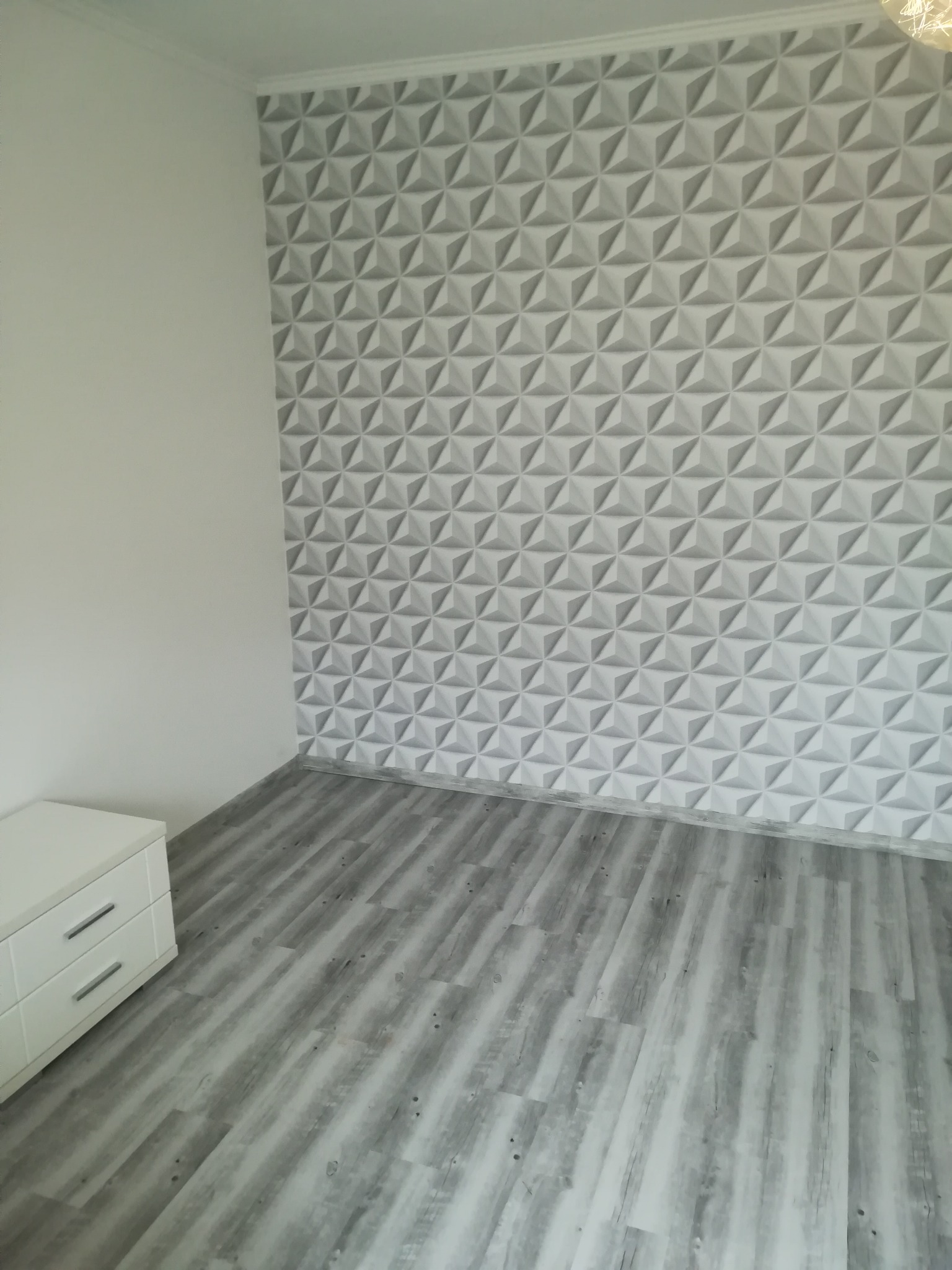 Pokój po remoncie z szarą podłogą z paneli imitujących drewno i ścianą z trójwymiarowych, geometrycznych paneli dekoracyjnych w odcieniach szarości i bieli.