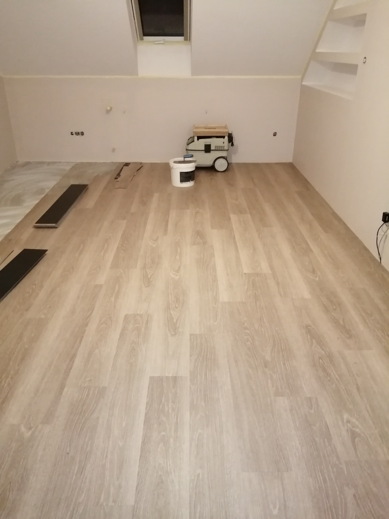 Jasna podłoga z paneli imitujących drewno, częściowo ułożona na poddaszu. W tle widoczny odkurzacz przemysłowy, wiadro z klejem i okno dachowe.
