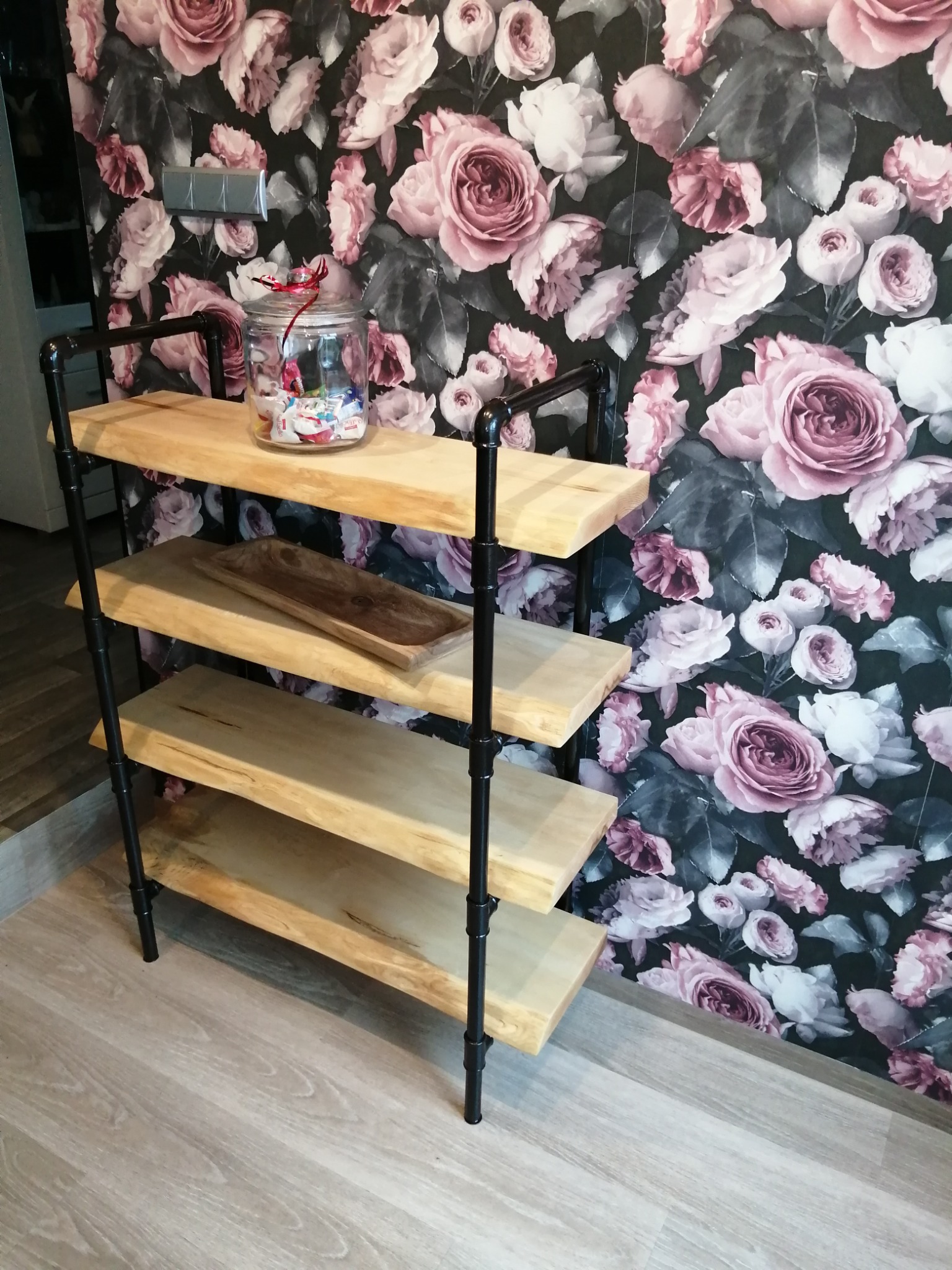 Ręcznie robiony regał z surowego drewna i czarnych rur, stojący na podłodze imitującej drewno, z tapetą w róże w tle. Na górnej półce słoik z zawartością, na drugiej drewniana misa.