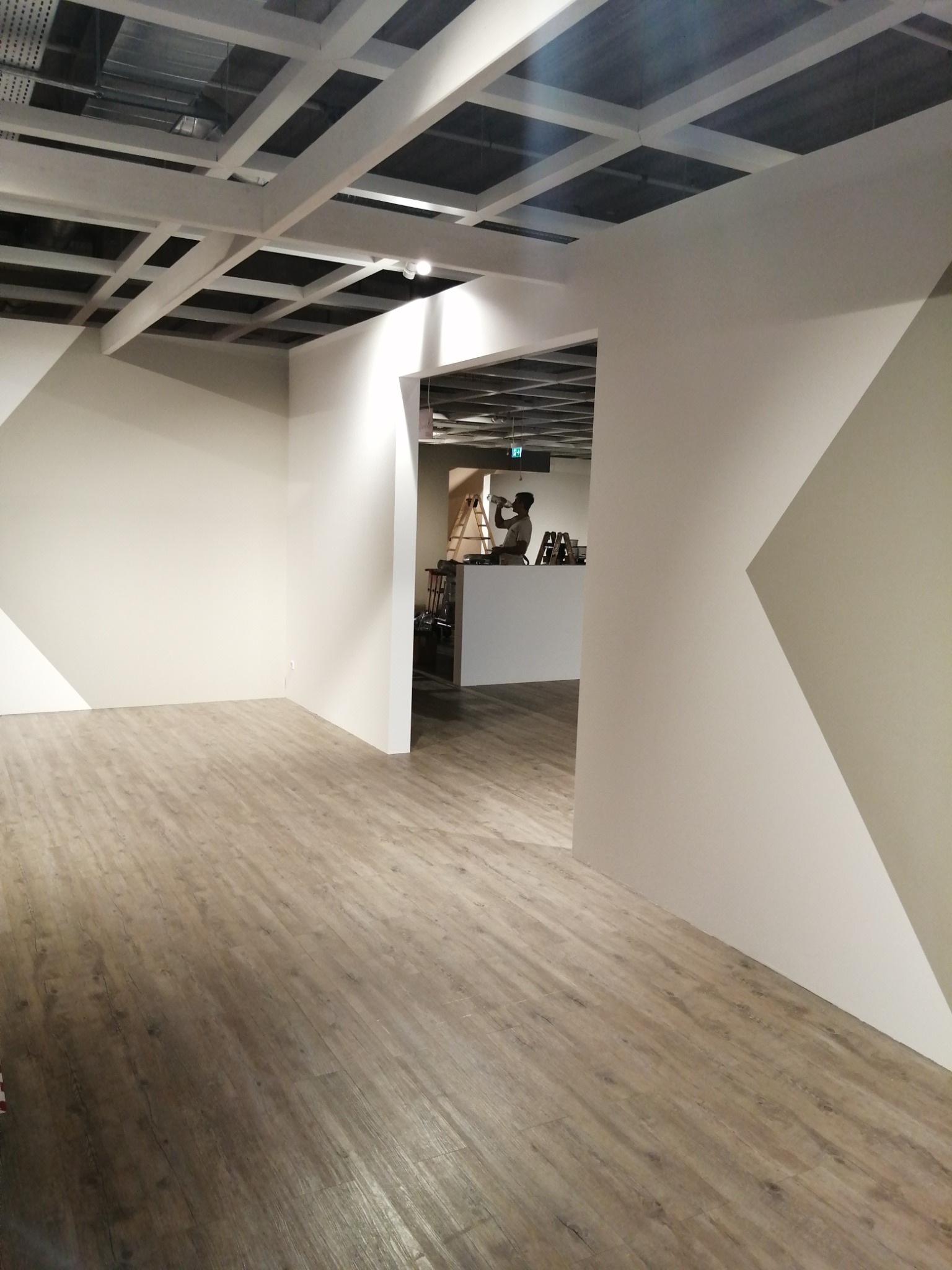 Wnętrze pomieszczenia z ułożoną podłogą z paneli imitujących drewno, widoczne ściany z geometrycznym wzorem i sufit z odkrytą konstrukcją, w tle mężczyzna pijący wodę na tle drabiny.