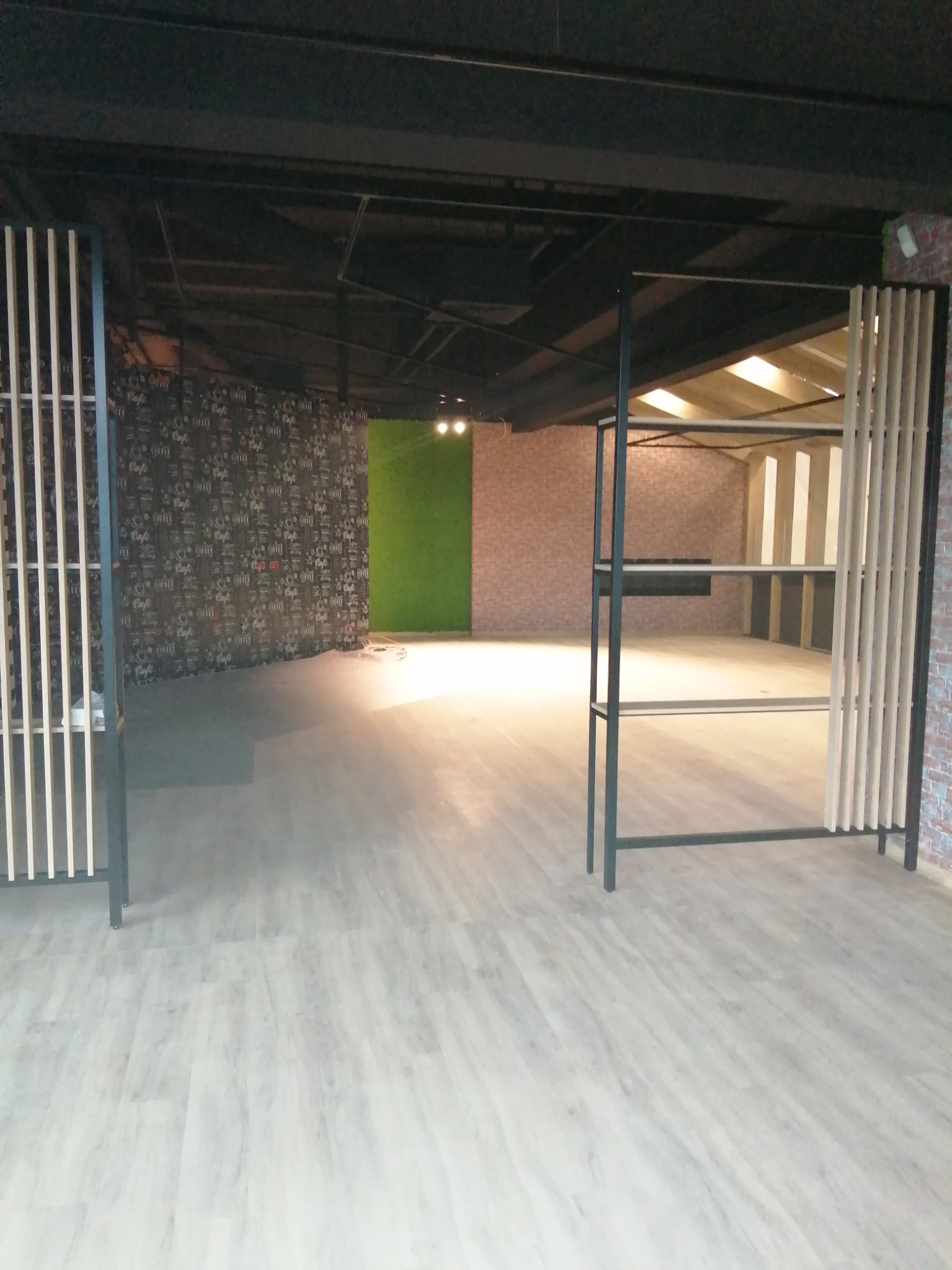 Surowe wnętrze lokalu usługowego z jasną podłogą imitującą drewno, czarnymi metalowymi konstrukcjami regałów i ścianami w różnych stylach: tapeta z motywem kawy, zielona ściana i cegła.