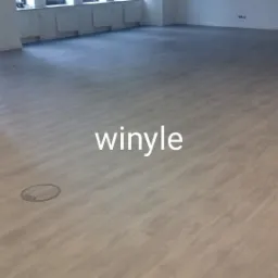 Jasnoszara podłoga winylowa imitująca drewno w przestronnym, jasnym pomieszczeniu biurowym z widocznymi włazami podłogowymi.