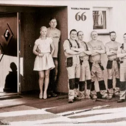 Grupa montażystów w kombinezonach pozuje z klientką przed wejściem do budynku z numerem 66, widoczna boczna część samochodu dostawczego.