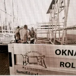 Montaż okien na budowie; pracownicy w trakcie pracy, w tle konstrukcja budynku, na pierwszym planie reklama firmy z hasłem '16 lat tradycji' i napisami 'Okna' i 'Role'.