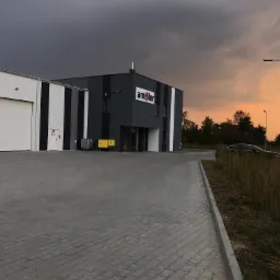 Nowoczesny budynek przemysłowy z bramą segmentową i logo firmy na fasadzie, widok z zewnątrz na tle zachodzącego słońca.