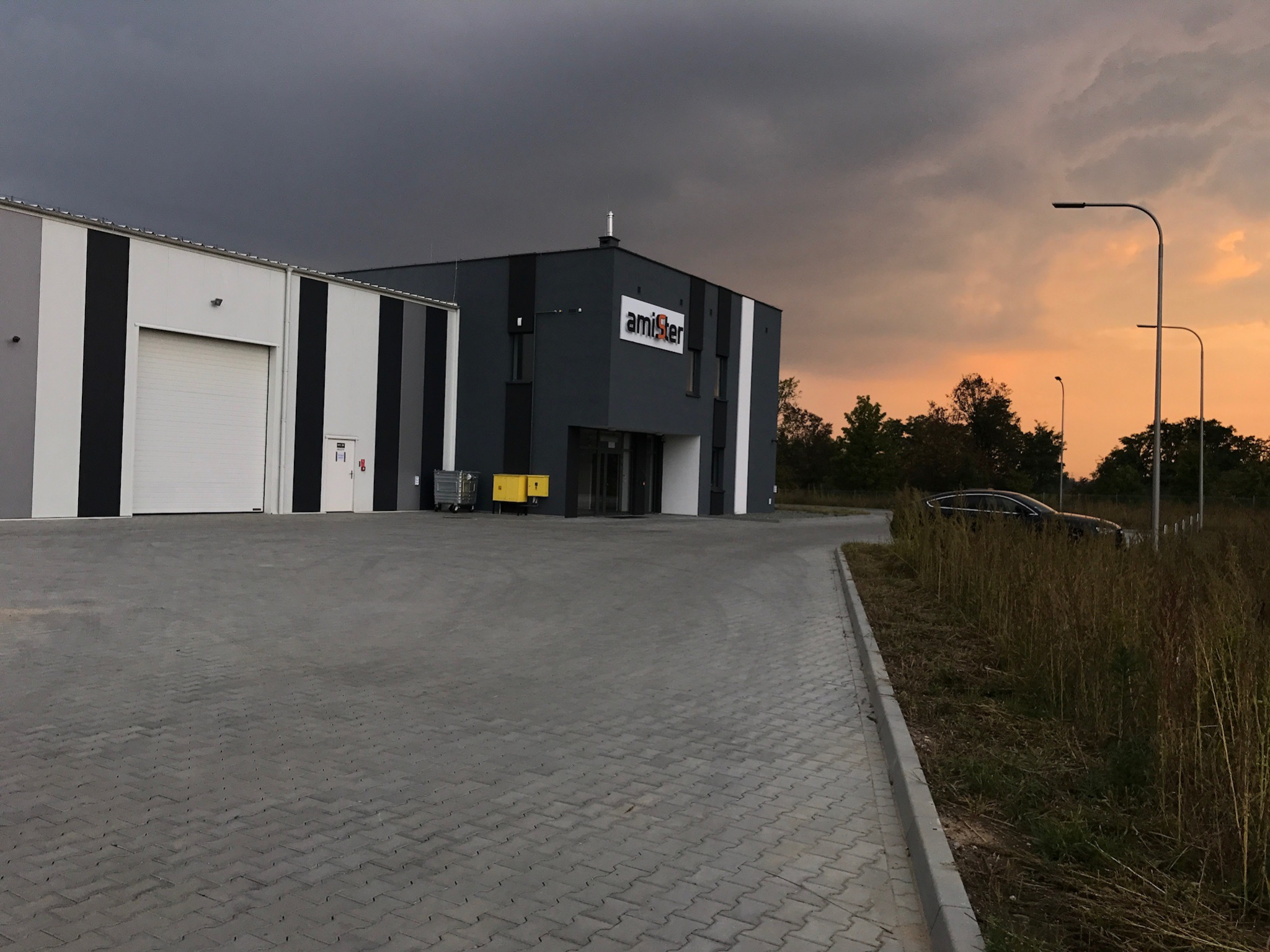 Nowoczesny budynek przemysłowy z bramą segmentową i logo firmy na fasadzie, widok z zewnątrz na tle zachodzącego słońca.