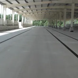 Wnętrze hali przemysłowej z betonową posadzką, widoczne stalowe słupy konstrukcyjne i kanały odwadniające w posadzce, ślady opon na posadzce.