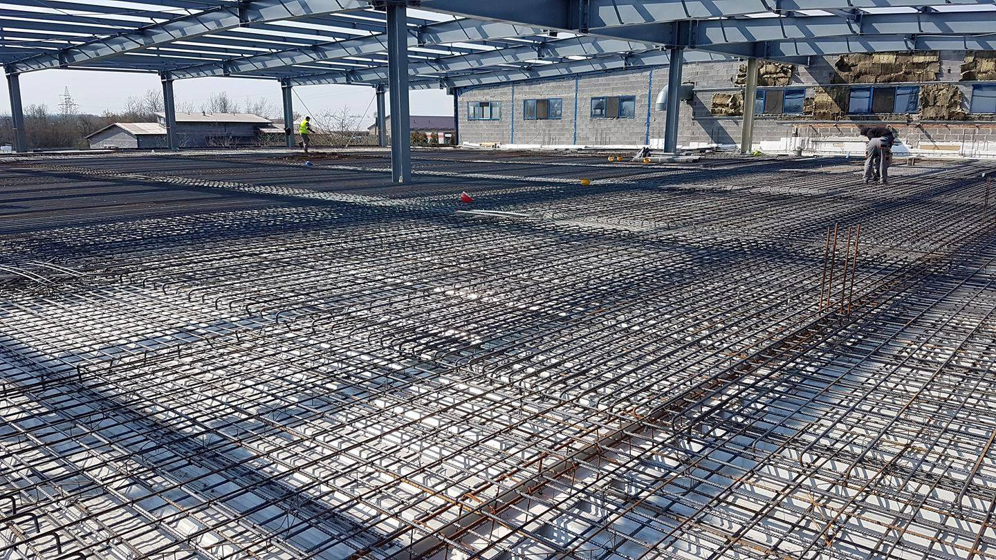 Siatka zbrojeniowa na dużej powierzchni w hali przemysłowej, widoczni pracownicy w oddali, konstrukcja stalowa dachu.