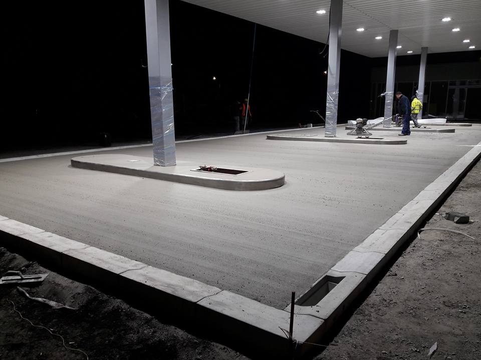 Świeżo wylana, szara posadzka na stacji benzynowej, wyrównywana przez pracownika za pomocą zacieraczki mechanicznej, z widocznymi słupami osłoniętymi folią.