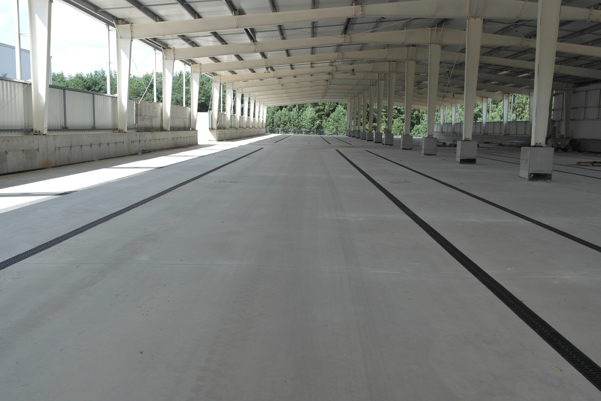 Wnętrze hali przemysłowej z betonową posadzką, widoczne stalowe słupy konstrukcyjne i kanały odwadniające w posadzce, ślady opon na posadzce.