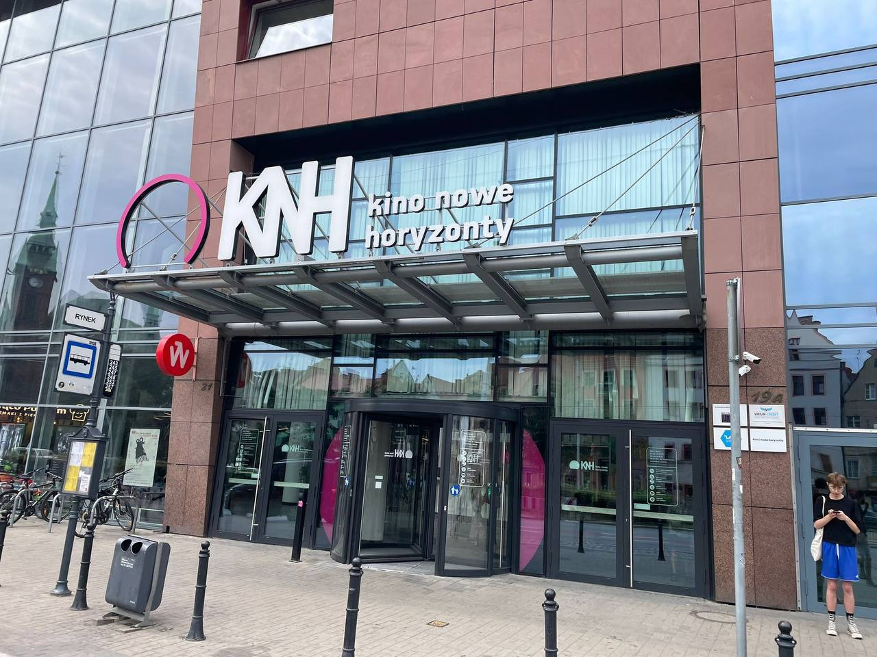 Fasada aluminiowo-szklana kina Nowe Horyzonty z obrotowymi drzwiami wejściowymi i dużymi przeszkleniami, widok z zewnątrz, w tle odbicie wieży kościoła.