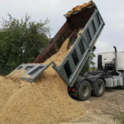 Ciężar&oacute;wka MAN wyładowuje piasek na placu budowy, widoczny fragment korony drzewa w tle.
