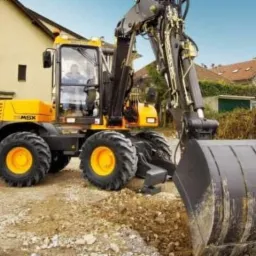 Żółto-czarna koparka kołowa MSX w trakcie prac ziemnych, z widocznym operatorem w kabinie i otoczeniem budynków mieszkalnych.