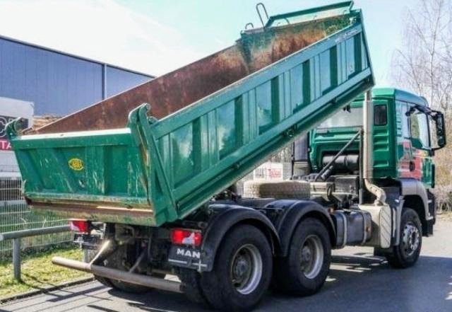 Zielona wywrotka MAN z podniesioną skrzynią ładunkową, widoczna z boku, na tle budynku i drzew.