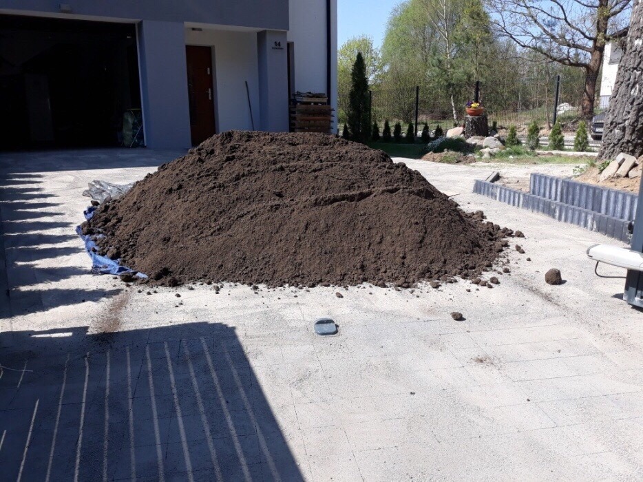 Kupa ciemnej ziemi na betonowej nawierzchni przed budynkiem z garażem, w tle widoczne drzewa i krzewy.