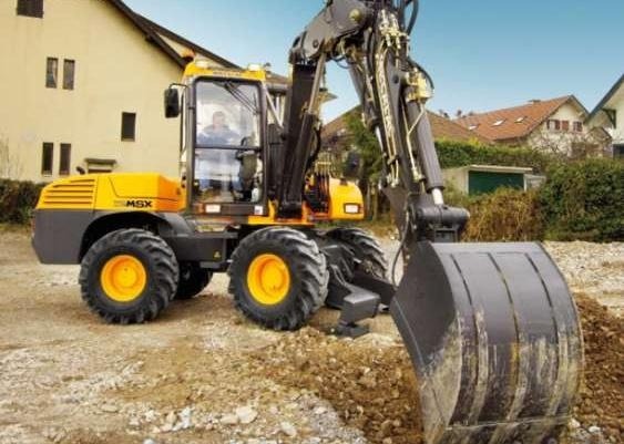 Żółto-czarna koparka kołowa MSX w trakcie prac ziemnych, z widocznym operatorem w kabinie i otoczeniem budynków mieszkalnych.