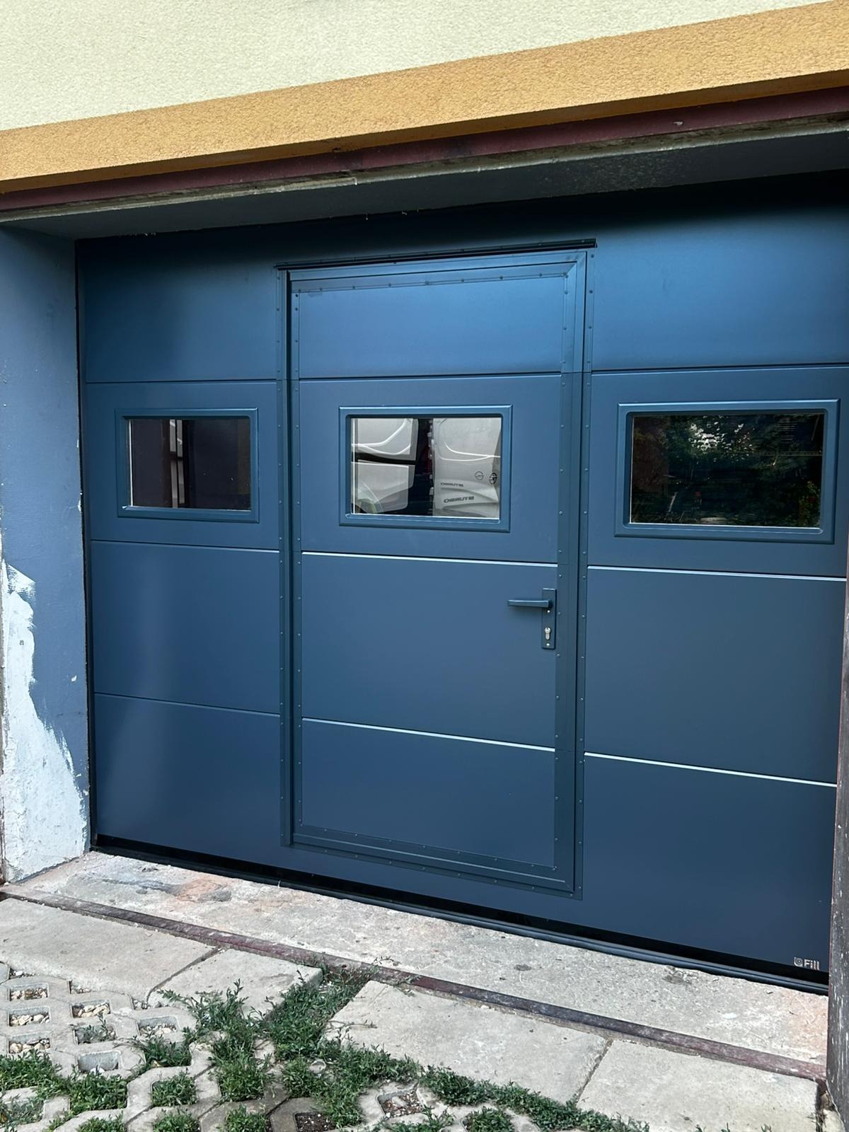 Nowoczesna, segmentowa brama garażowa w odcieniach szarości z trzema małymi oknami i drzwiami wejściowymi, zamontowana w garażu.