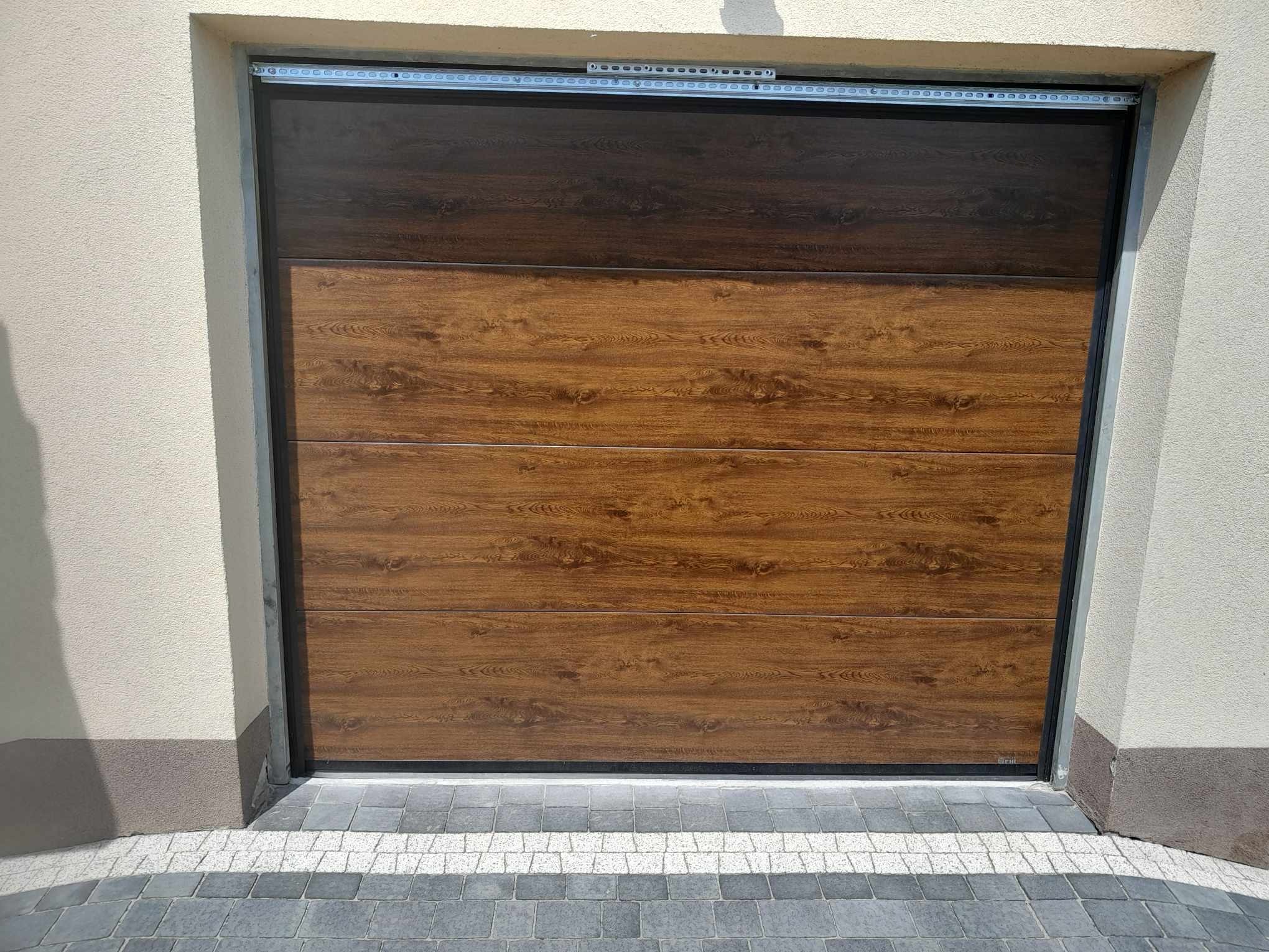 Zamknięta, segmentowa brama garażowa w odcieniach brązu, imitująca drewno. Widoczne prowadnice i fragment elewacji budynku. Kostka brukowa u dołu kadru.