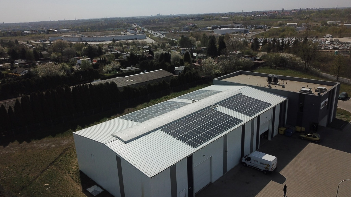 Widok z góry na magazyn z panelami słonecznymi na dachu, z widocznym fragmentem miasta w tle.