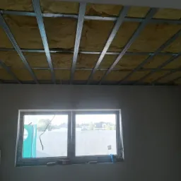 Poddasze w trakcie ocieplania wełną mineralną, widoczne metalowe profile pod zabudowę, okno z widokiem na domy jednorodzinne.