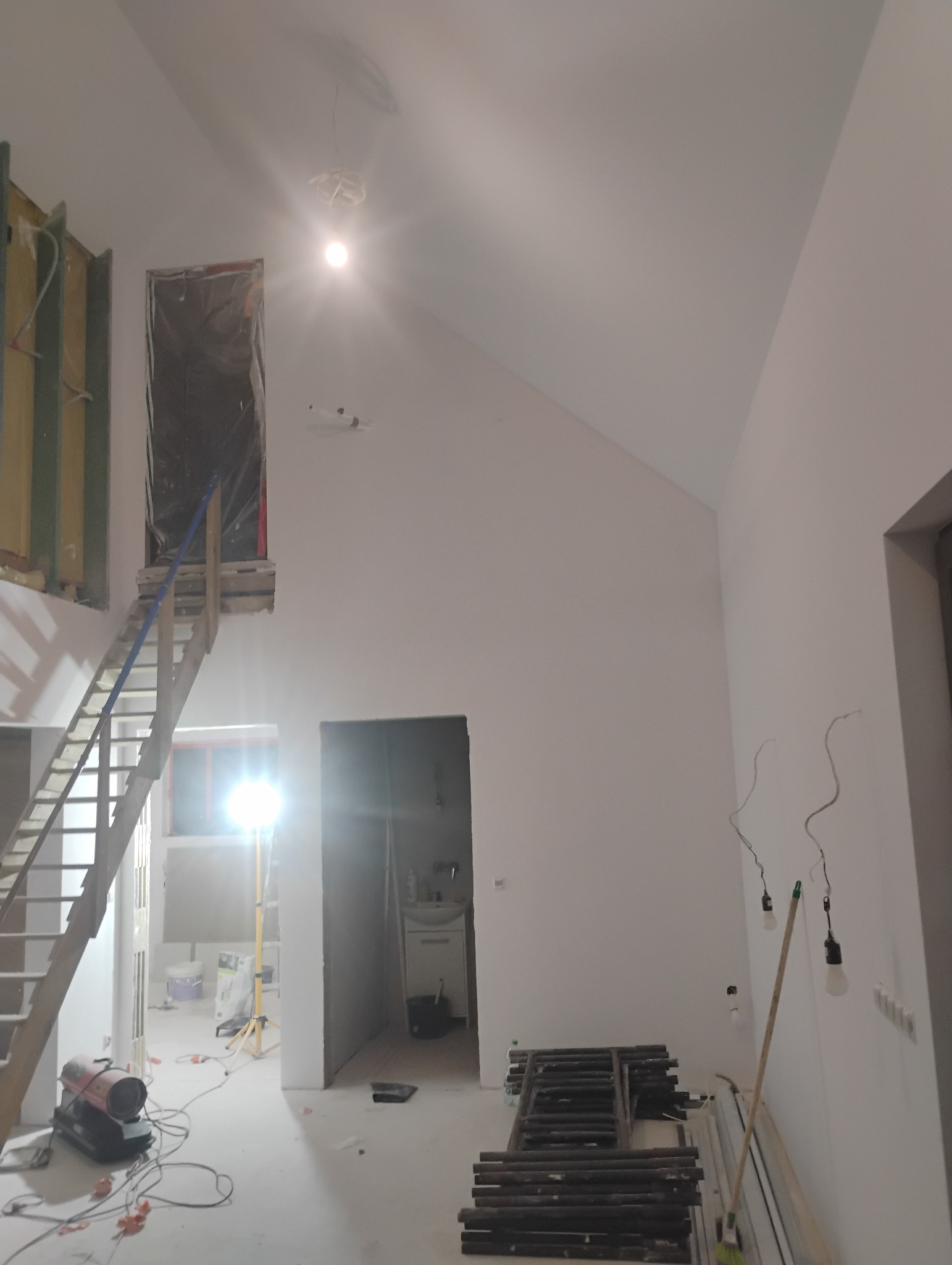 Wysokie wnętrze w trakcie prac wykończeniowych: białe ściany, drewniane schody prowadzące na antresolę, widoczne rusztowania i lampy budowlane. W tle okno z ciepłym światłem.