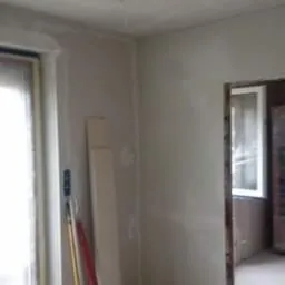 Pokój w trakcie prac wykończeniowych: ściany po nałożeniu gładzi, widoczne narzędzia, butelka i niedokończone wykończenie przy oknie.