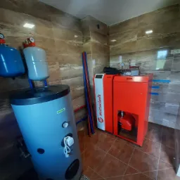 Wnętrze kotłowni z piecem na paliwo stałe Heitztechnik, zbiornikiem buforowym i instalacją wodną, widoczne rury miedziane i elementy sterujące.