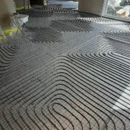 Rozłożone rury ogrzewania podłogowego na folii z nadrukiem w surowym wnętrzu domu w trakcie budowy, widok na sąsiednie pomieszczenie i okno.