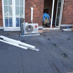 RAKSO Oskar Olender - Montaż klimatyzacji na dachu budynku z czerwonej cegły; widoczny agregat LG, butla z gazem i instalator w niebieskiej koszuli w otwartym oknie.