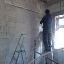 RAKSO Oskar Olender - Instalacja przewodów elektrycznych w surowym wnętrzu z białych bloczków, widoczny monter na rusztowaniu i drabina