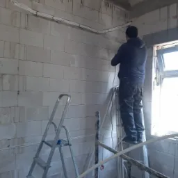 Instalacja przewodów elektrycznych w surowym wnętrzu z białych bloczków, widoczny monter na rusztowaniu i drabina