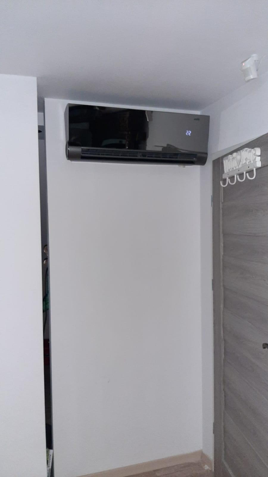 Czarny klimatyzator ścienny z wyświetlaczem temperatury 22 stopnie Celsjusza, zamontowany w rogu pokoju obok szarej drewnopodobnej futryny drzwi z wieszakami.