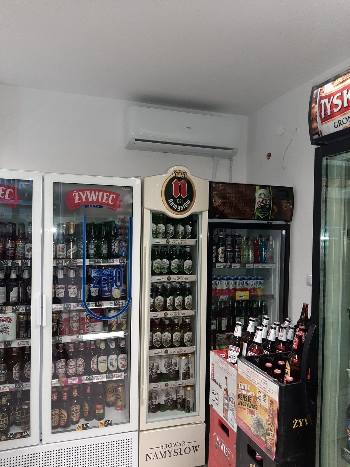 Biały klimatyzator ścienny zamontowany nad chłodziarkami z piwem różnych marek w sklepie, widoczne butelki Żywca i Namysłowa.