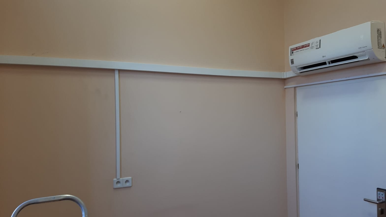Biały klimatyzator LG zamontowany na ścianie w pokoju, widoczne listwy maskujące przewody i gniazdko elektryczne, fragment drabiny w lewym dolnym rogu.