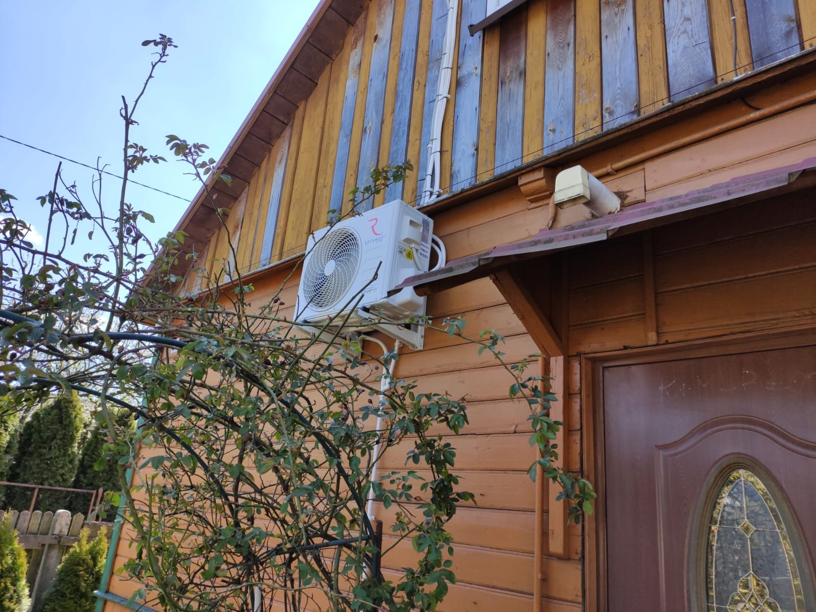 Biała jednostka zewnętrzna klimatyzacji zamontowana na pomarańczowej, drewnianej ścianie domu, widoczna spod gałęzi krzewu róż z młodymi liśćmi.